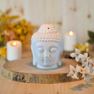 Pode incluir: Um difusor de óleo com cabeça de Buda em cerâmica branca, com olhos fechados e expressão serena. O difusor está sobre uma fatia de madeira, com velas acesas e flores ao fundo. A cabeça de Buda tem cerca de 15 cm de altura.