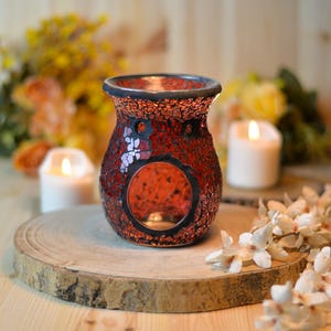 Peut inclure: Un diffuseur d'huiles essentielles en verre mosaïque rouge avec une ouverture ronde et un bord noir. Le diffuseur est posé sur une tranche de bois avec des bougies allumées et des fleurs en arrière-plan. Le motif en mosaïque est fait de petits morceaux de verre rouge irréguliers.