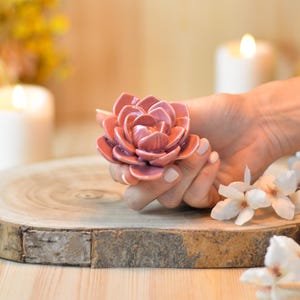 Può includere: Una scultura floreale in ceramica nei toni del rosa e malva, tenuta in mano. Il fiore è dettagliato con petali a strati. La scultura è su una superficie di legno, con uno sfondo sfocato di candele e fiori.