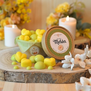 Puede incluir: Una exhibición de madera presenta ceras perfumadas en forma de corazón en tonos amarillos y verdes, junto a un recipiente redondo etiquetado "Melón". Las ceras están dispuestas en una pequeña cesta y dispersas sobre una superficie de madera, con velas encendidas y flores al fondo.