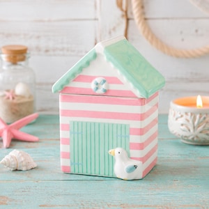 Può includere: Un barattolo in ceramica a forma di cabina da spiaggia con coperchio rimovibile. La struttura presenta strisce orizzontali rosa e bianche, un tetto verde menta e una porta verde menta. Una piccola figura di gabbiano bianco è fissata sulla base anteriore.
