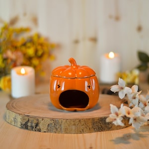 Può includere: Un bruciatore di olio in ceramica arancione a forma di zucca con ritagli a stella e coperchio. Il bruciatore è su una fetta di legno con candele accese e fiori sullo sfondo. Un oggetto decorativo per la stagione autunnale.