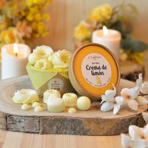 Può includere: Esposizione di cere profumate al limone in varie forme, tra cui ciambelle e sfere, presentate su una fetta di legno. Un contenitore rotondo aperto con la scritta "Crema de Limón" è visibile. La scena è illuminata da candele accese e decorazioni floreali.
