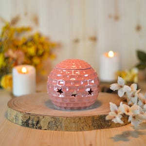 May include: A pink ceramic sphere-shaped essential oil diffuser with a textured surface and star cutouts. The diffuser sits on a wooden slice with lit candles and flowers in the background, creating a warm, inviting ambiance.