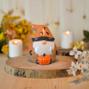 Può includere: Una statuetta di gnomo in ceramica con un cappello arancione e una barba bianca, che tiene in mano una piccola zucca arancione. Lo gnomo è marrone e bianco con dettagli neri. La statuetta è su una fetta di legno, con candele accese e fiori sullo sfondo.