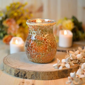May include: A mosaic glass candle holder in orange, gold, and green hues. The candle holder is displayed on a wooden slice, with a backdrop of flowers and lit candles. The overall effect is a warm and inviting atmosphere.