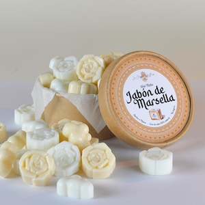 Puede incluir: Una colección de ceras perfumadas de color marfil en varias formas florales, que se derraman de una caja de papel kraft. Un recipiente redondo de cartón con el texto "Jabón de Marsella" está abierto junto a la caja. Las ceras están hechas de cera de soja 100% natural.