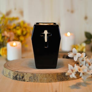 Puede incluir: Un difusor de aceites esenciales de cerámica negra en forma de ataúd con un corte de cruz blanco. El difusor se encuentra sobre una rodaja de madera, con velas encendidas y flores en el fondo.