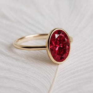May include: A gold ring featuring a large, oval-cut ruby gemstone. The ruby is a vibrant red color and is set in a bezel setting. The ring's band is a simple, polished gold. The ring is displayed on a white, feathery surface.