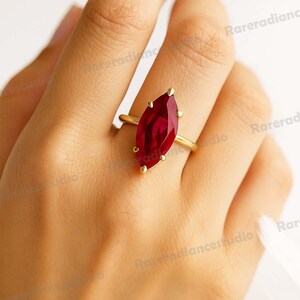 May include: A gold ring with a marquise-cut, deep red gemstone. The ring has a thin band, and the gemstone is secured by four prongs. The ring is displayed on a person's finger.