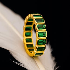 Op de afbeelding: Een gouden ring met rechthoekige smaragdgroene stenen. De ring ligt op een witte veer tegen een zwarte achtergrond. Het ontwerp toont een reeks smaragdgeslepen stenen, die een doorlopende band van kleur vormen.