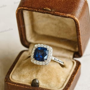 Può includere: Un anello d'argento con una pietra preziosa quadrata blu scuro, circondata da pietre più piccole e chiare. L'anello è esposto in un portagioie in pelle marrone con interno beige. La scatola è rettangolare con coperchio incernierato.
