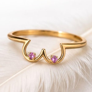 May include: A gold-colored ring with a unique design featuring two pink gemstones. The ring's shape is a stylized representation of a pair of breasts. The ring is set on a white feather.