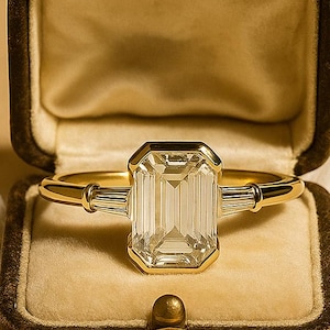 May include: A gold ring with a large emerald-cut diamond center stone and two smaller baguette diamonds on either side. The ring is displayed in an open, vintage-style brown jewelry box with a cream-colored interior.