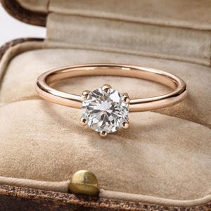Peut inclure: Une bague de fiançailles en or rose avec un gros diamant clair rond serti dans une monture à six griffes. La bague est présentée dans un écrin à bijoux vintage en cuir marron avec un intérieur beige.