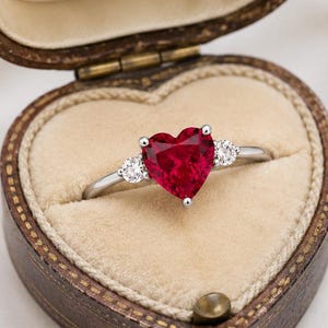 May include: A silver-toned ring featuring a large, heart-shaped red gemstone at the center, flanked by two smaller, round, clear stones on each side. The ring is displayed inside an open, heart-shaped, dark brown, vintage-style velvet box.