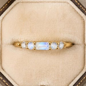 May include: A gold ring featuring a central rectangular baguette-cut moonstone flanked by two smaller round moonstones and two tiny round clear stones on each side, all set in a thin gold band and presented inside an octagonal velvet ring box.