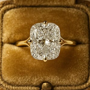 Könnte beinhalten: Ein goldener Ring mit einem großen, rechteckig geschliffenen Diamanten, gefasst in einem goldenen Band. Der Ring wird in einer samtigen, aufklappbaren Schatulle in warmem Goldbraun präsentiert. Der Diamant funkelt mit vielen Facetten.