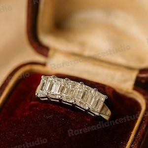 Puede incluir: Anillo de platino con cinco diamantes talla esmeralda, dispuestos en fila. El anillo se exhibe en un joyero vintage de terciopelo rojo con cierre dorado. El diseño del anillo es elegante y clásico.