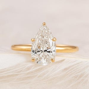 May include: A gold engagement ring with a pear-shaped diamond. The diamond is clear and faceted, set on a thin gold band. The ring is displayed on a white, textured surface.