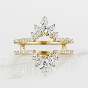 May include: A gold-colored ring with a double band design. The top band features a row of small, clear stones, while the center has a cluster of larger, marquise-cut stones. The ring is set against a white background.