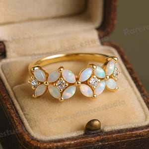 May include: A gold ring with a floral design, set with opals and diamonds. The ring is displayed in a brown jewellery box with a velvet lining. The opal stones are oval and arranged to look like flowers. The ring's band is slender.