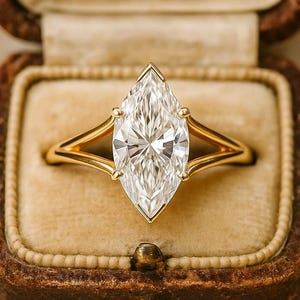 Op de afbeelding: Een verlovingsring met een marquise-geslepen diamant en een gouden band, gepresenteerd in een sieradendoosje in vintage-stijl. De diamant is groot en helder, met een briljante slijpvorm. De gouden band heeft een split-schacht ontwerp.