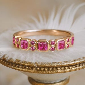 May include: A gold ring featuring a series of square and round pink gemstones. The ring is set on a white surface with a gold-colored decorative base. The gemstones are a vibrant pink color, and the ring has a polished finish.
