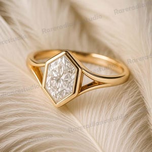 Puede incluir: Un anillo de oro con una piedra central hexagonal en forma de diamante. El anillo tiene un diseño de caña dividida y está colocado sobre un fondo blanco suave.