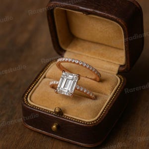 May include: A rose gold engagement ring set with an emerald-cut diamond and a matching band, displayed in a vintage brown leather ring box with a tan velvet interior. The rings are adorned with small, sparkling accent stones.