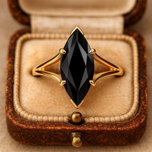 以下が含まれることがあります： 大きなマーキス型の黒い宝石が付いたゴールドリング。リングは、繊細な透かし模様が施されたゴールドバンドにセットされています。茶色のヴィンテージスタイルのジュエリーボックスに、ベージュの内装で展示されています。
