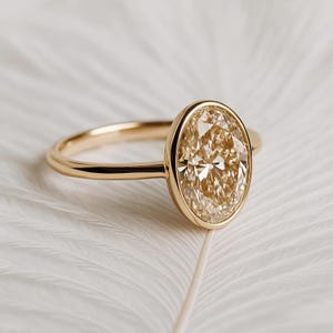 May include: A gold ring with a large, oval-cut diamond. The diamond has a warm, champagne color and is set in a bezel setting. The band is a simple, polished gold. The ring is displayed on a white surface.