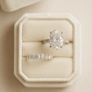 Puede incluir: Dos anillos de diamantes en un joyero de terciopelo color crema. El anillo superior presenta un gran diamante de talla ovalada con una banda pavimentada. El segundo anillo es una banda con múltiples diamantes redondos. La caja está abierta.