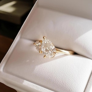 May include: A gold ring featuring two pear-cut diamonds. The ring is set in a gold band and presented in a white box. The diamonds are clear and catch the light, creating a sparkling effect.