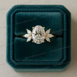 May include: A gold ring with a large oval-cut diamond center stone, flanked by six smaller marquise-cut diamonds. The ring is set in a gold band and is displayed in a teal velvet ring box.