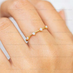 May include: A delicate gold ring with five small, round, white opal gemstones. The ring is set on a slender gold band, creating a minimalist and elegant design. The ring is shown on a person's finger.