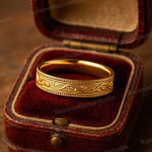 Może przedstawiać: Złoty pierścionek z grawerowanym wzorem kwiatowym, prezentowany w czerwonym aksamitnym pudełku. Pierścionek ma szczegółowy wzór wokół obrączki. Otwarte pudełko ukazuje misterne detale pierścionka.