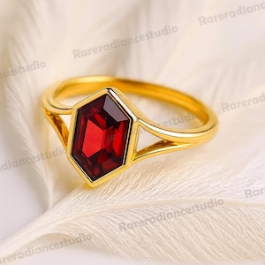 May include: A gold ring with a hexagonal, deep red gemstone. The ring has a delicate band and a geometric design, set against a soft, white background. The ring's design is modern and elegant.
