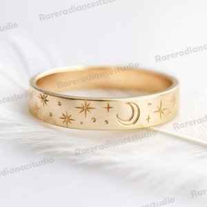 Puede incluir: Un anillo dorado con un diseño celestial grabado. El anillo presenta estrellas, una luna creciente y pequeños puntos. El anillo está sobre un fondo blanco.