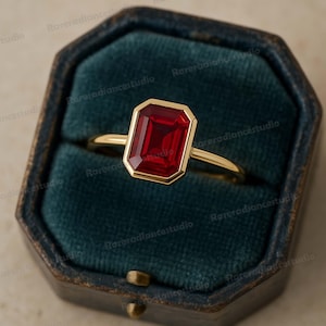 Könnte beinhalten: Ein goldener Ring mit einem rechteckigen, tiefroten Edelstein. Der Edelstein ist in einer goldenen Lünette gefasst, und der Ring wird in einer dunkelblauen Schmuckschatulle präsentiert. Der Ring hat ein dünnes, goldenes Band.
