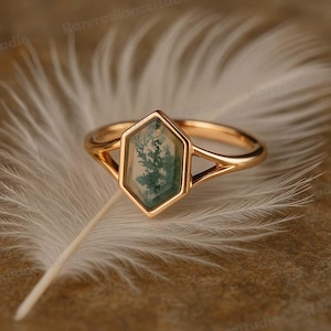 May include: A gold ring with a hexagonal, moss agate stone. The stone features green and white patterns resembling a miniature landscape. The ring's band has a split design, adding a unique touch to the jewelry.