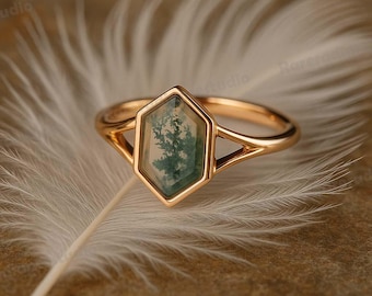 May include: A gold ring with a hexagonal, moss agate stone. The stone features green and white patterns resembling a miniature landscape. The ring's band has a split design, adding a unique touch to the jewelry.