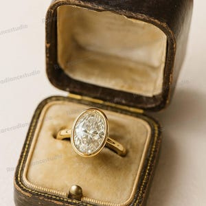 Könnte beinhalten: Ein goldener Ring mit einem oval geschliffenen Diamanten in einer Zargenfassung, präsentiert in einer offenen, braunen Schmuckschatulle aus Leder im Vintage-Stil mit beigem Innenraum. Der Ring hat einen schmalen, polierten Ring.