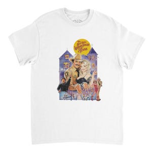May include: White t-shirt featuring a colorful graphic from the movie "The Best Little Whorehouse in Texas." The design includes a yellow sun, a blue house, and characters from the film. The text "The Best Little Whorehouse in Texas" is also visible.
