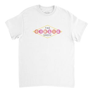 Puede incluir: Camiseta blanca con un gráfico en forma de diamante. El gráfico presenta el texto "THE GIRLIE SHOW" en un estilo retro, con la palabra "GIRLIE" escrita en círculos coloridos. La camiseta está hecha de un material suave y cómodo.