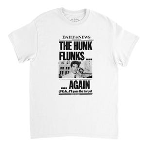 May include: White t-shirt featuring a black and white graphic resembling a newspaper headline. The headline reads "THE HUNK FLUNKS... AGAIN" with a photo of two men. Text at the bottom says "JFK Jr.: I'll pass the bar yet".