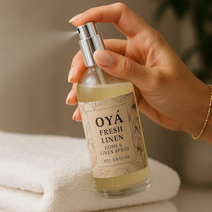 May include: A clear glass spray bottle labeled "OYÁ FRESH LINEN HOME & LINEN SPRAY NET 3.4 FL OZ" is held in a hand, spraying a fine mist. The bottle sits above a stack of white and tan towels.