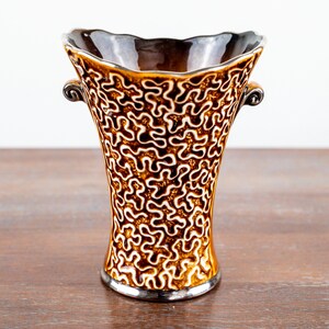 Peut inclure: Un vase en céramique avec un bord évasé et festonné et deux petites anses en forme de volute. L'extérieur est orné d'un motif en relief blanc rappelant un labyrinthe sur un vernis brun brillant, tandis que l'intérieur est brun foncé.