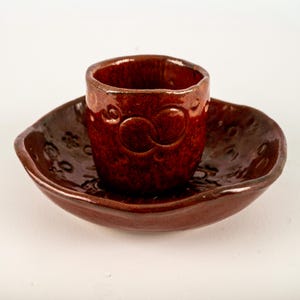 May include: A handmade, dark brown ceramic cup and saucer set. The cup has a rounded shape with embossed details, including a pair of circles. The saucer is oval-shaped with floral patterns. The set is glazed with a glossy finish.