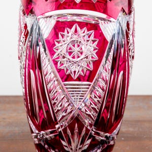 Puede incluir: Un jarrón de cristal rojo rubí con intrincados patrones geométricos y un diseño de estrella. El jarrón tiene un borde festoneado y un cuerpo facetado, mostrando la artesanía. Es un artículo decorativo.